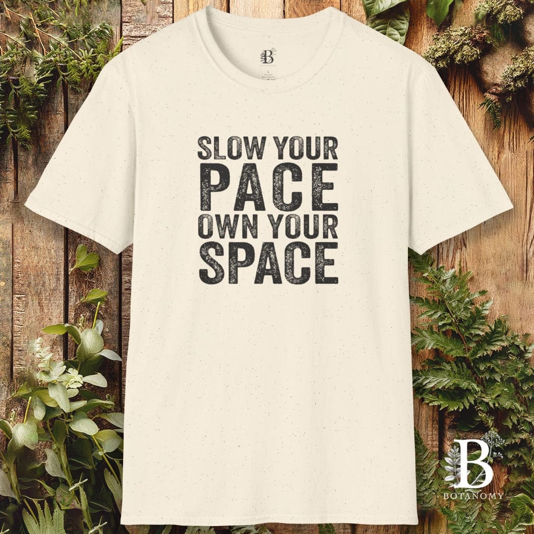 Beige t-shirt with 'Slow your pace, own your space' text on a wooden background with plants.