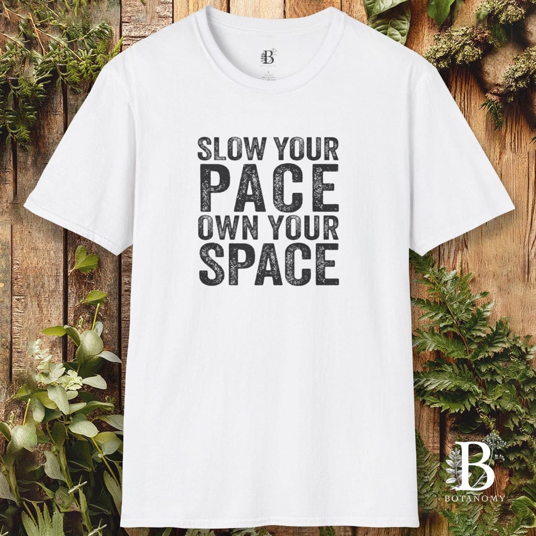 White t-shirt with 'Slow your pace, own your space' text on a wooden background with plants.