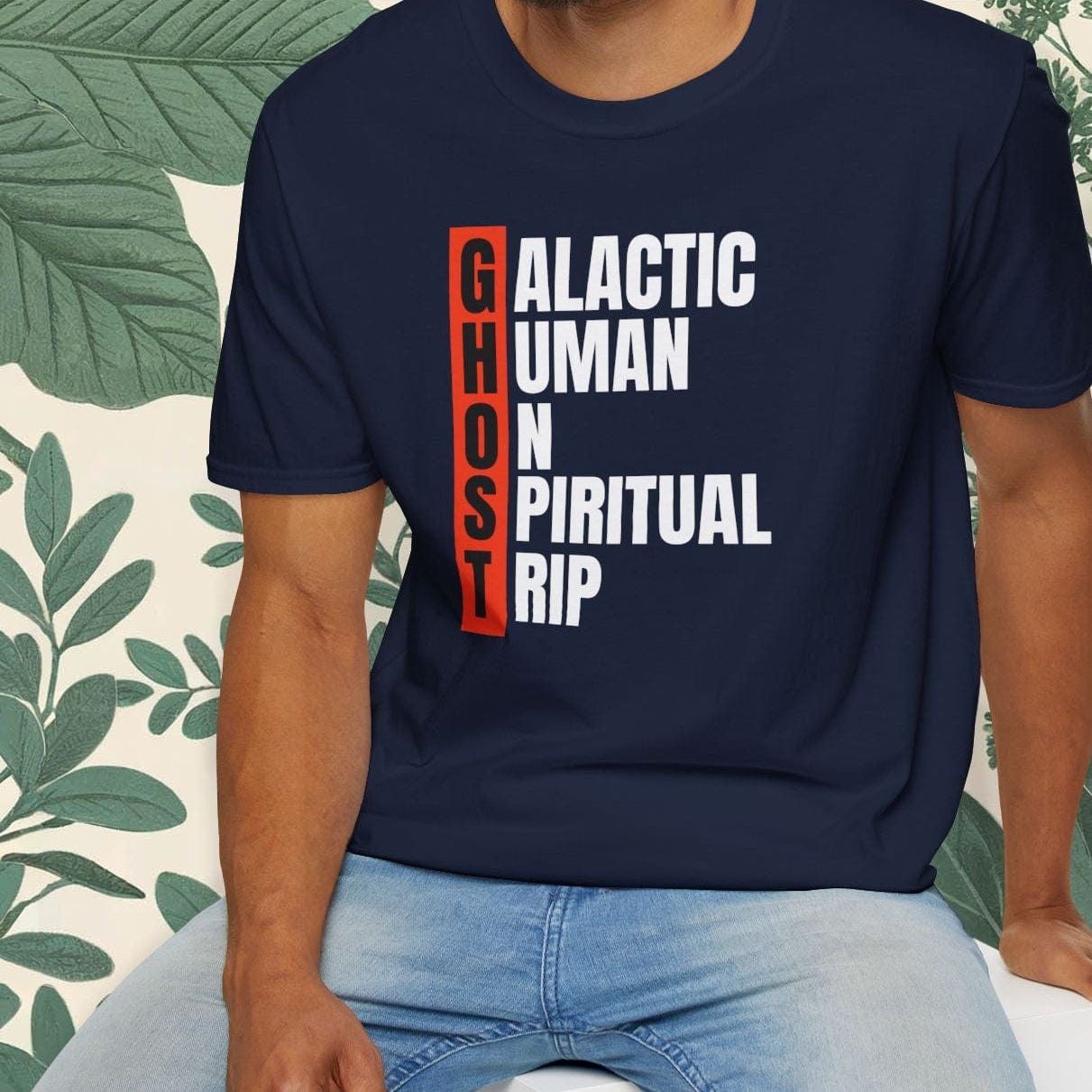 Man wearing a navy blue t-shirt with text design, sitting on a white stool with a green leafy background.