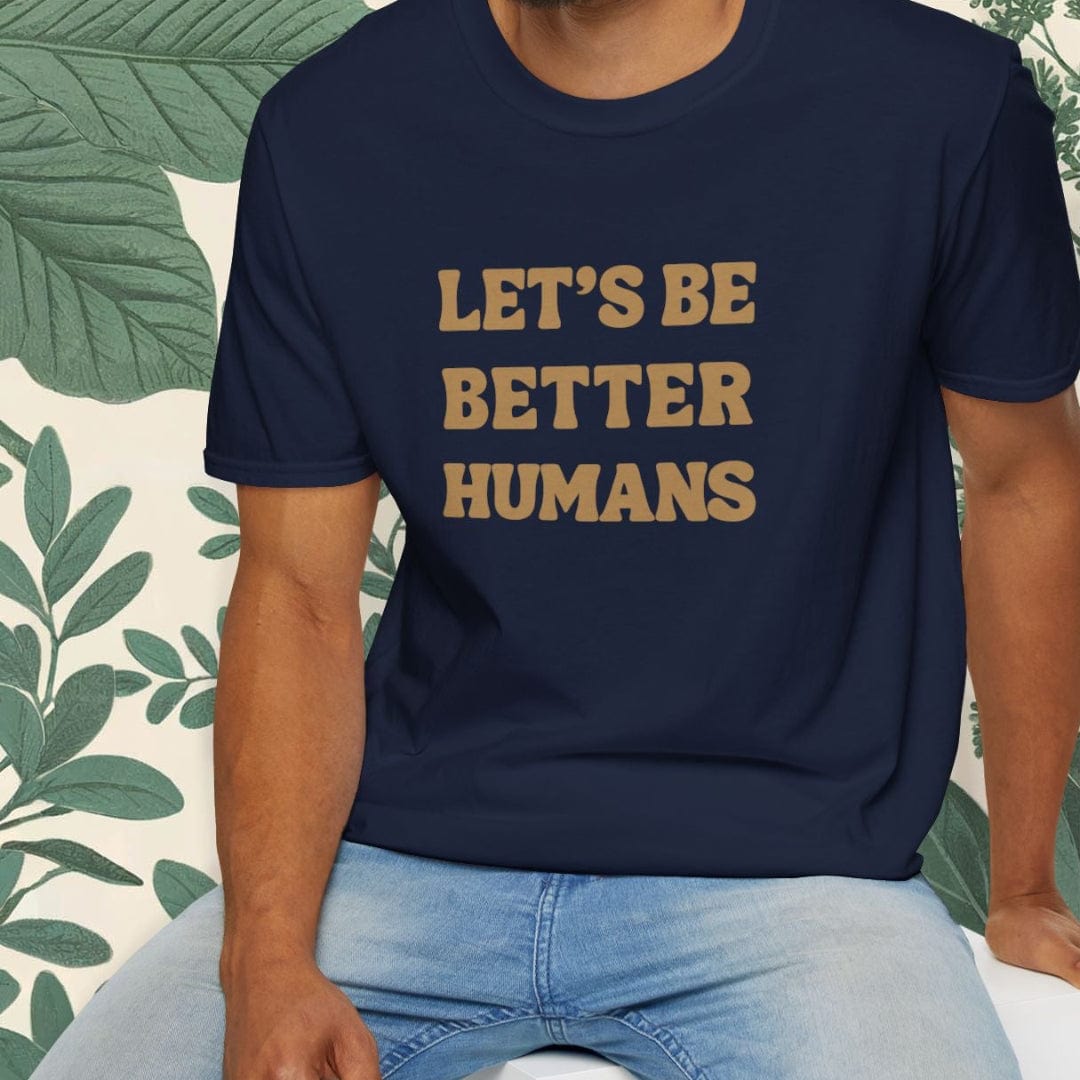 Man wearing a navy blue t-shirt with 'LET'S BE BETTER HUMANS' text, sitting on a white stool with a green leafy background.