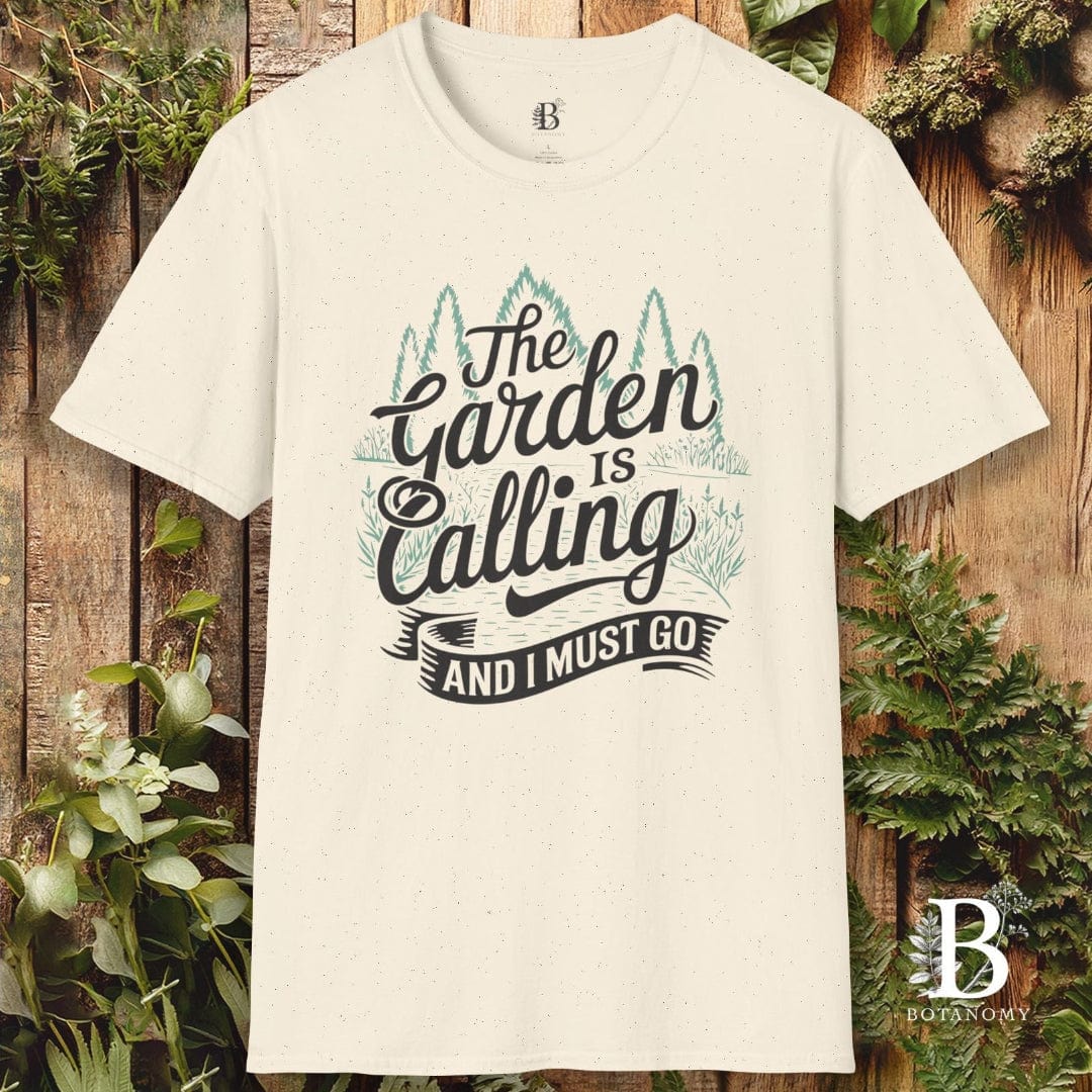 White t-shirt with 'The Garden is Calling and I Must Go' text on a wooden background with plants.