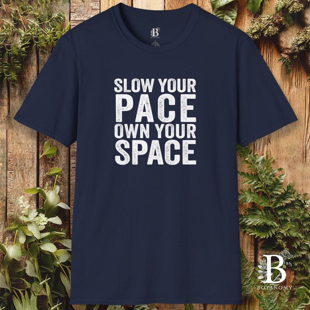 Navy blue t-shirt with white text 'Slow your pace, own your space' on a wooden background with plants.