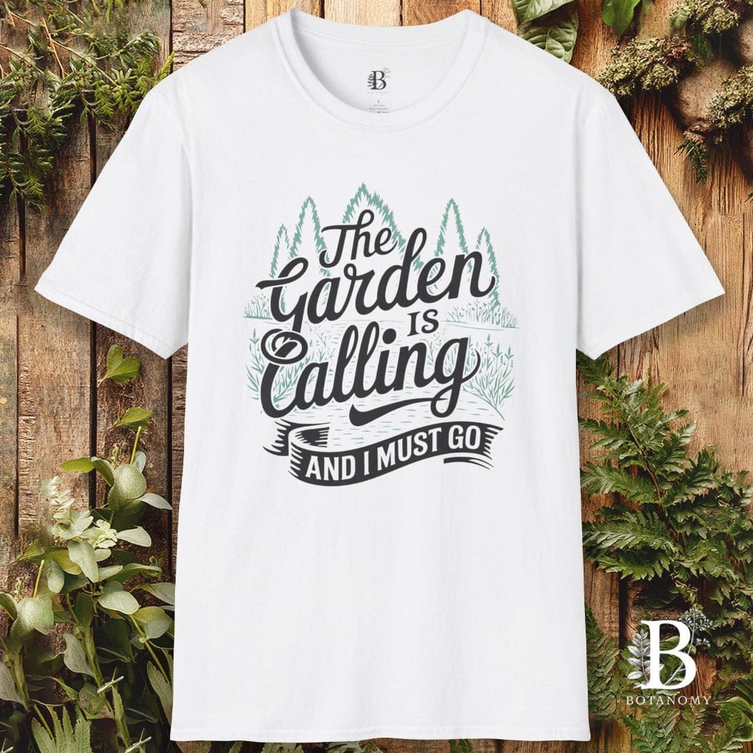 White t-shirt with 'The Garden is Calling and I Must Go' text on a wooden background with plants.
