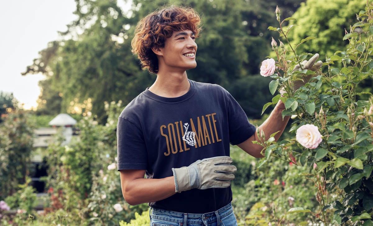 Man in a garden wearing a 'Soil mate' t-shirt and gardening gloves, surrounded by plants and flowers.