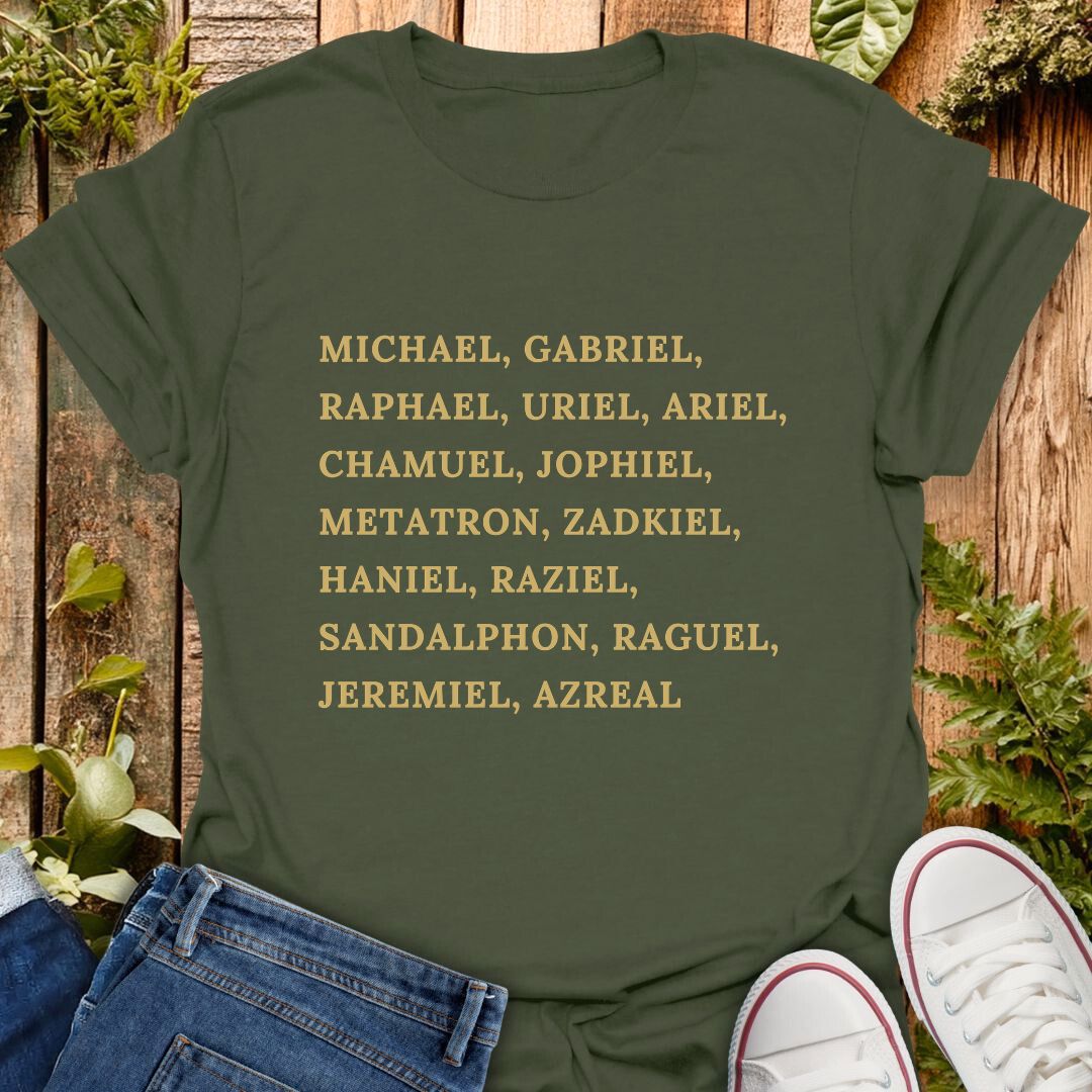An olive green short-sleeve T-shirt laid flat on a rustic wooden surface with greenery and jeans, with a pair of white sneakers partly visible at the bottom right. The shirt has a list of names of archangels printed in gold text of on the front.