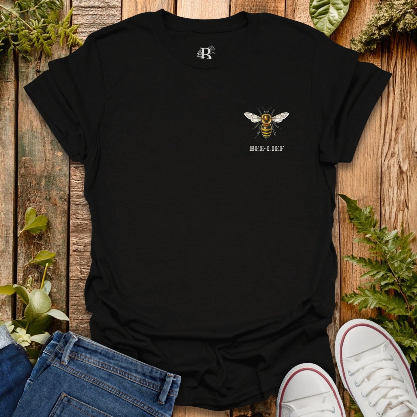 Black t-shirt with a bee design and text on a wooden surface with plants.