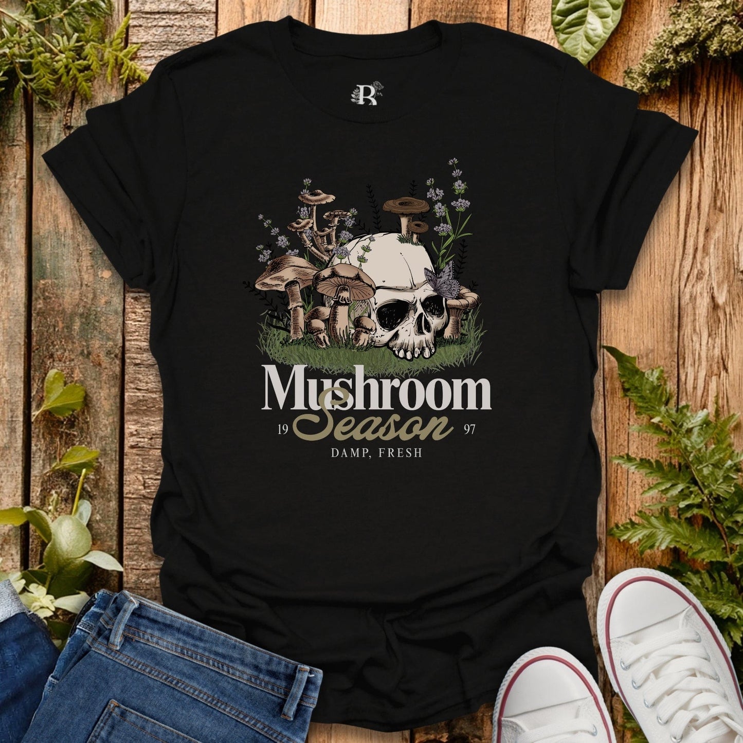 Black t-shirt with mushroom and skull design on a wooden surface with plants.