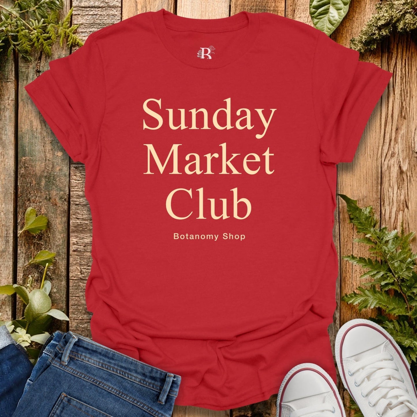 Red t-shirt with 'Sunday Market Club' text on a wooden surface with plants.