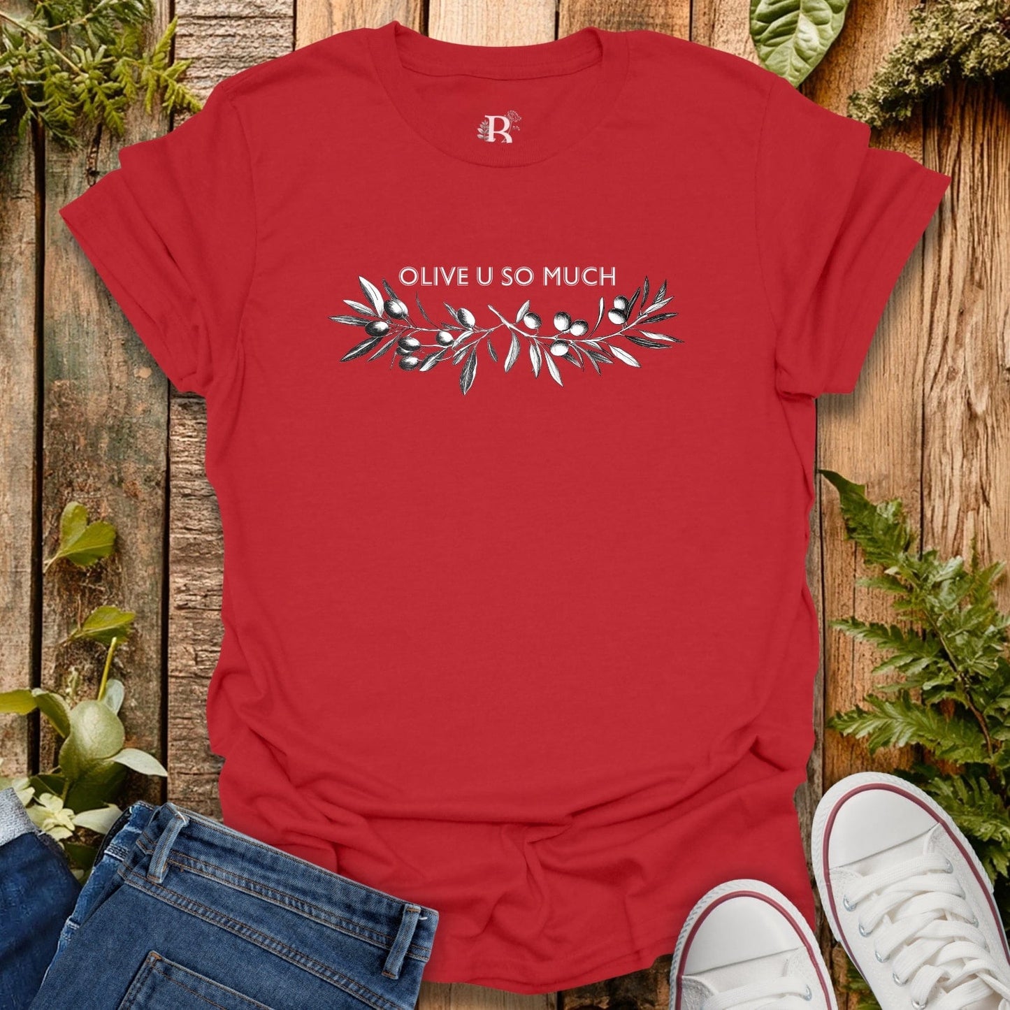 Red t-shirt with 'OLIVE U SO MUCH' design on a wooden surface with plants.