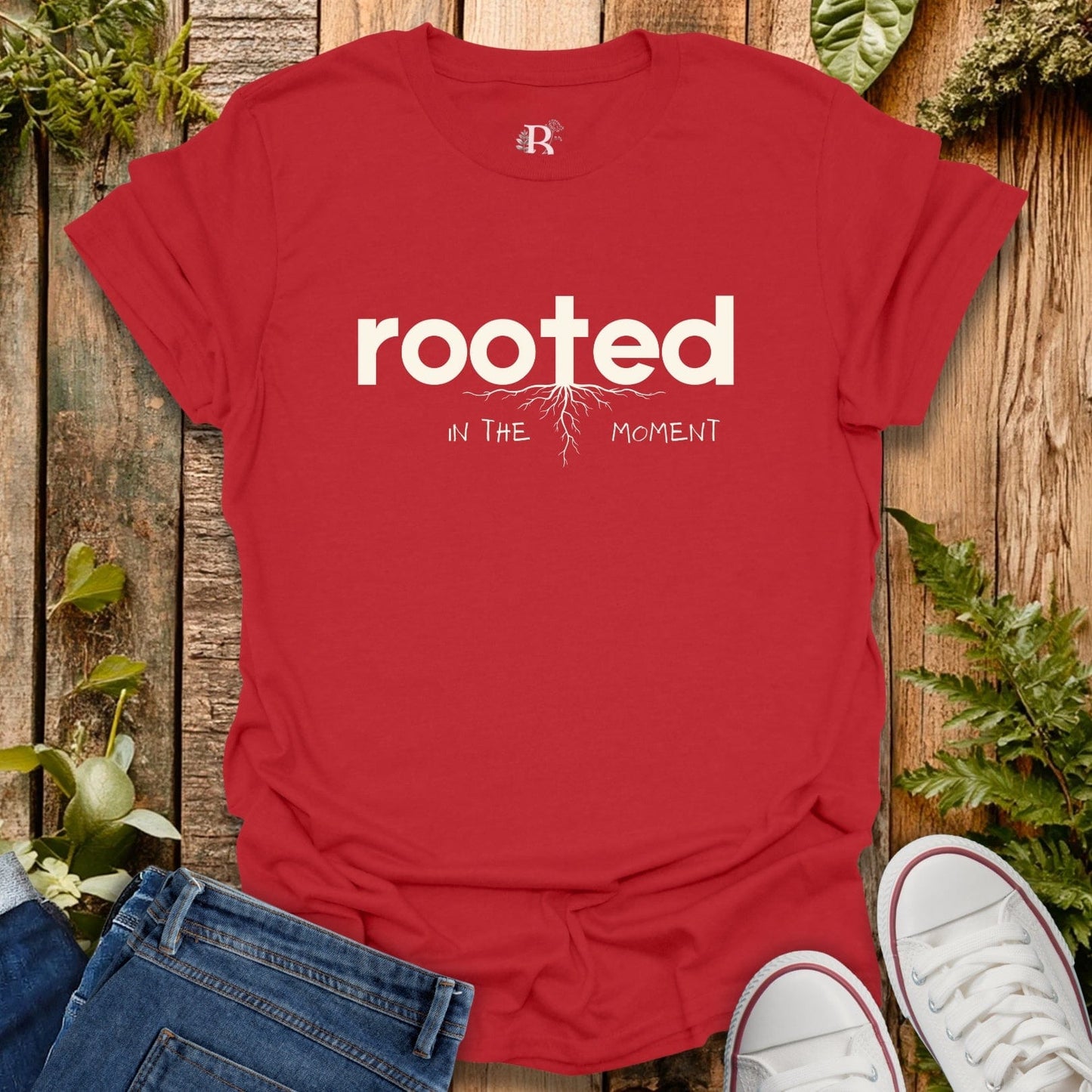 Red t-shirt with 'rooted in the moment' text on a wooden surface with plants.