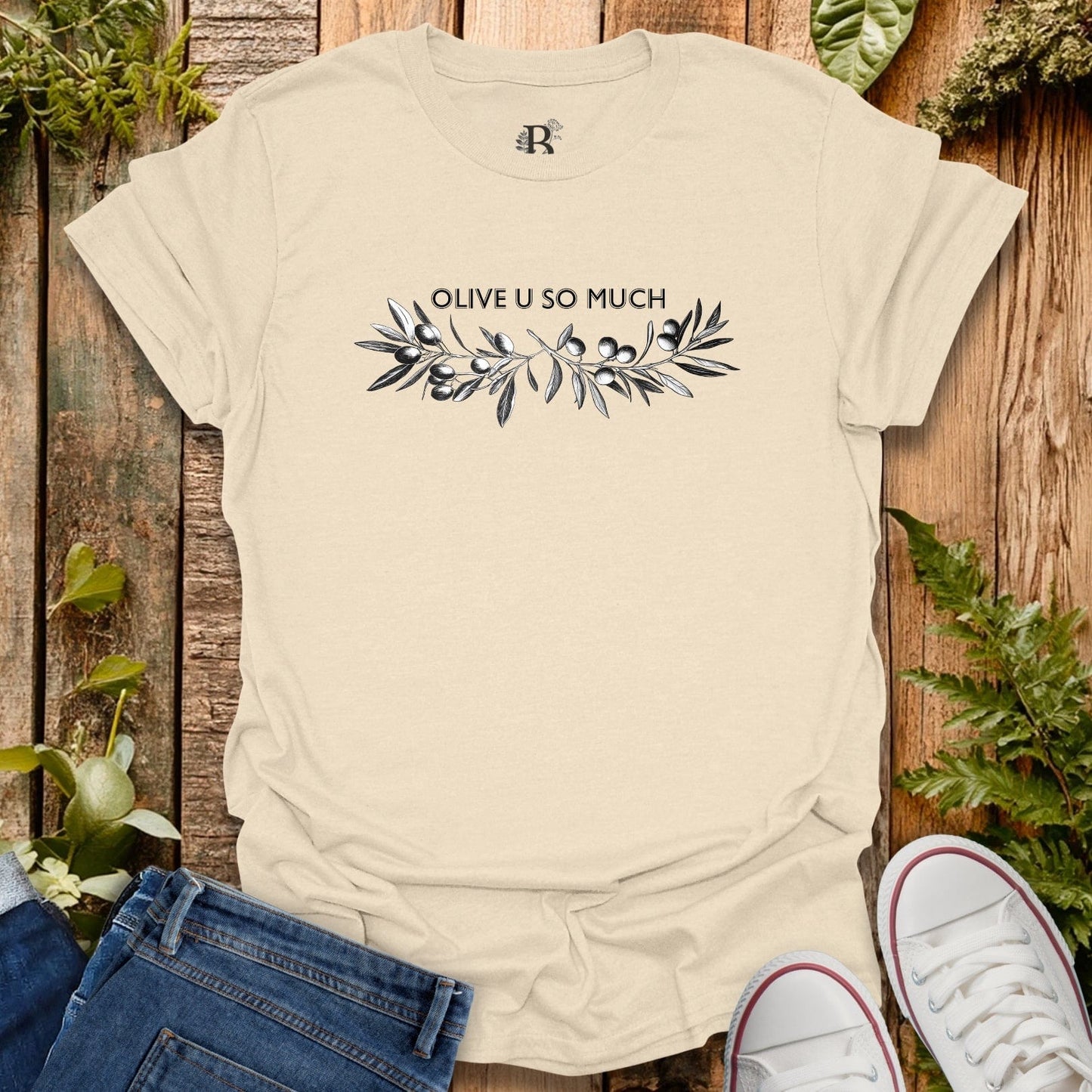 Beige t-shirt with 'OLIVE U SO MUCH' text and leaf design on a wooden background with plants.