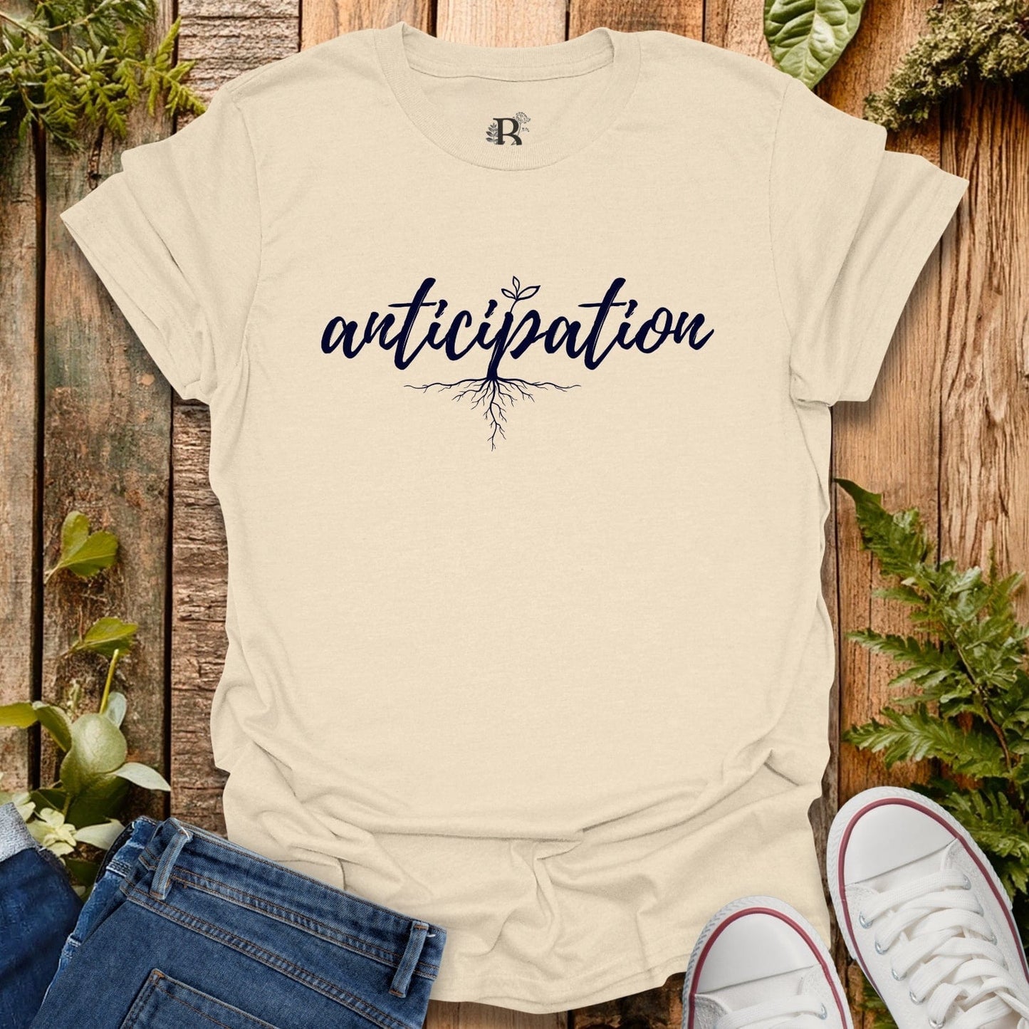 Beige t-shirt with 'anticipation' text on a wooden background with plants.