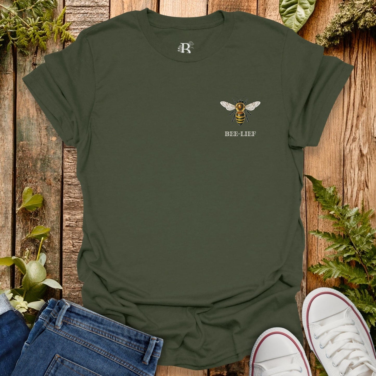 Green t-shirt with a bee design and 'BEE-LIEF' text on a wooden surface with plants.