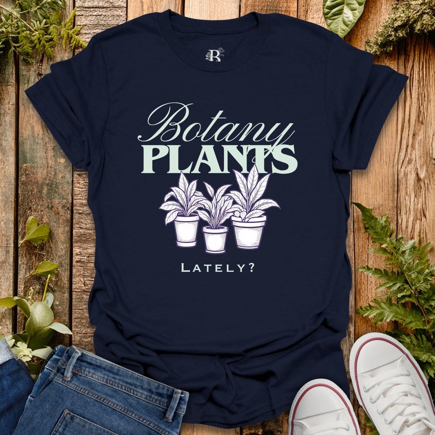 Navy blue t-shirt with 'Botany Plants Lately?' design on a wooden surface with plants.