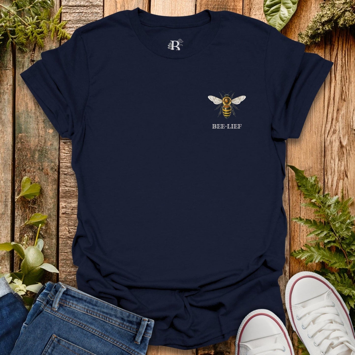 Navy blue t-shirt with bee design and 'BEE-LIEF' text on a wooden surface with plants.