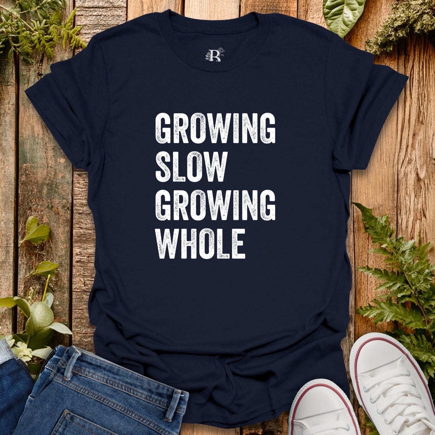 Navy blue t-shirt with 'GROWING SLOW GROWING WHOLE' text on a wooden surface with plants.