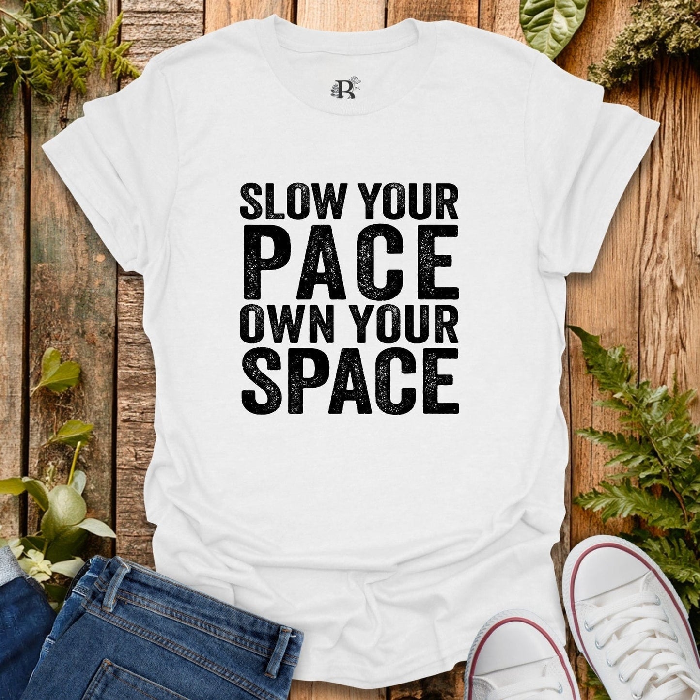 White t-shirt with 'Slow your pace, own your space' text on a wooden background