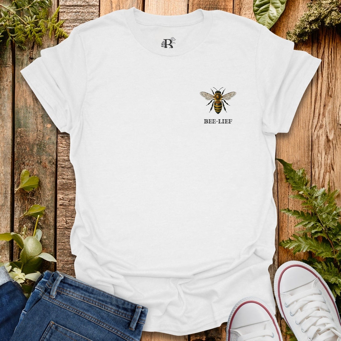 White t-shirt with bee graphic and 'BEE-LIEF' text on a wooden surface with plants.