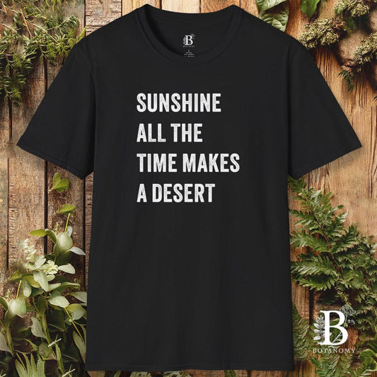 Black t-shirt with white text 'Sunshine all the time makes a desert' on a wooden background with plants.