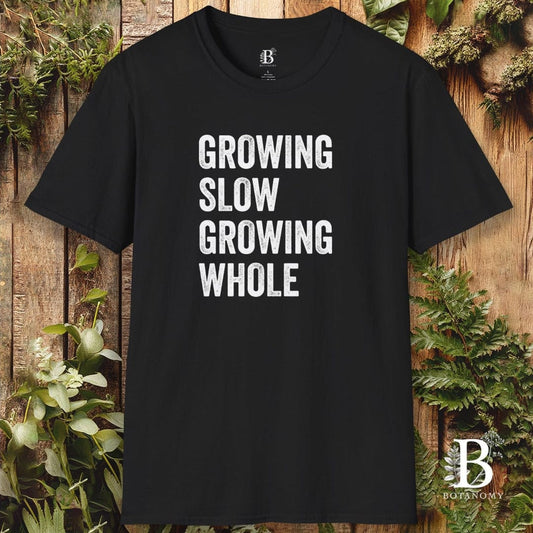 Black t-shirt with 'GROWING SLOW GROWING WHOLE' text on a wooden background with plants.