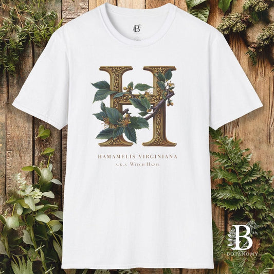White t-shirt with a decorative letter 'H' design on a wooden background with greenery