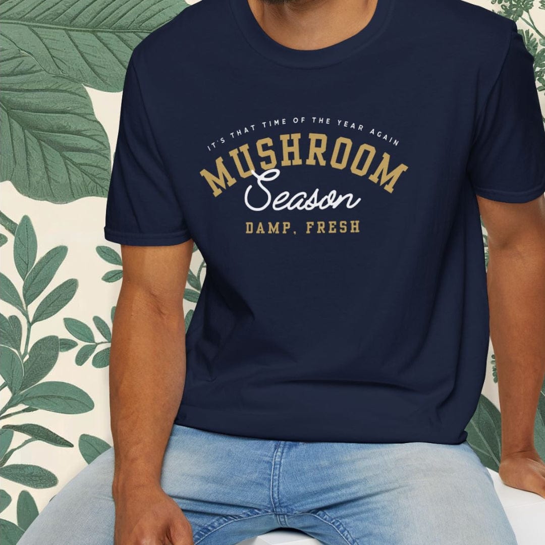 Man wearing a navy blue t-shirt with text, sitting on a white stool against a white background with green leafy elements.