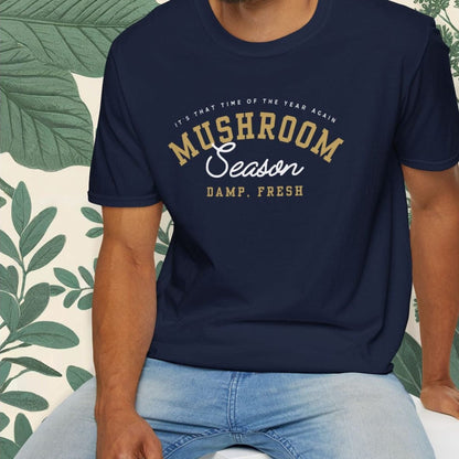 Man wearing a navy blue t-shirt with text, sitting on a white stool against a white background with green leafy elements.