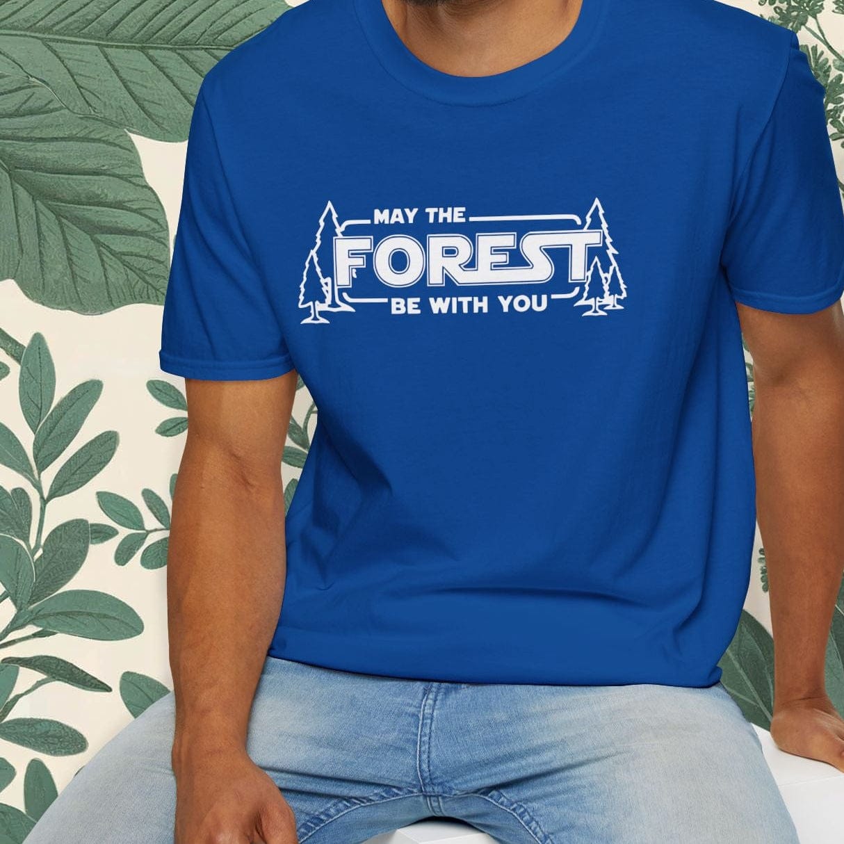 Man wearing a blue t-shirt with 'May the Forest Be With You' text, sitting on a white stool with greenery in the background.