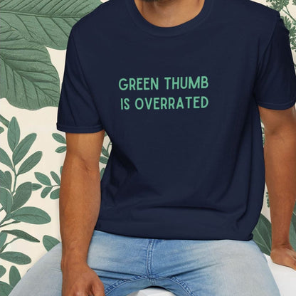 Man wearing a navy blue t-shirt with 'Green Thumb is Overrated' text, sitting on a white stool with a plant-themed background.