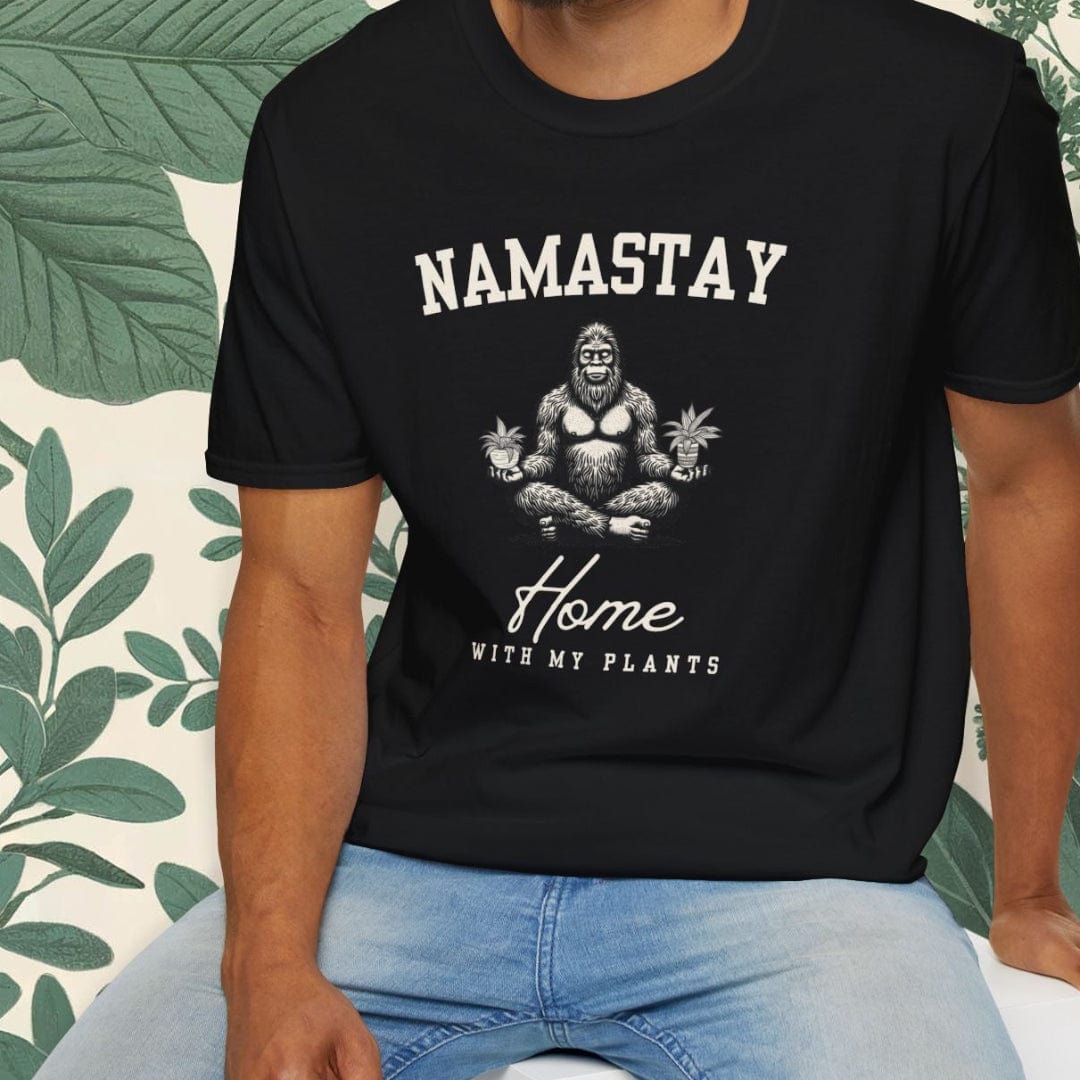 Man wearing a black t-shirt with 'NAMASTAY Home With My Plants' design, sitting on a white stool with a green leafy background.