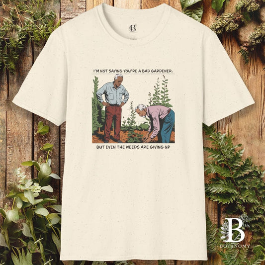 Beige t-shirt with a graphic of two men gardening and text, against a wooden background with plants.