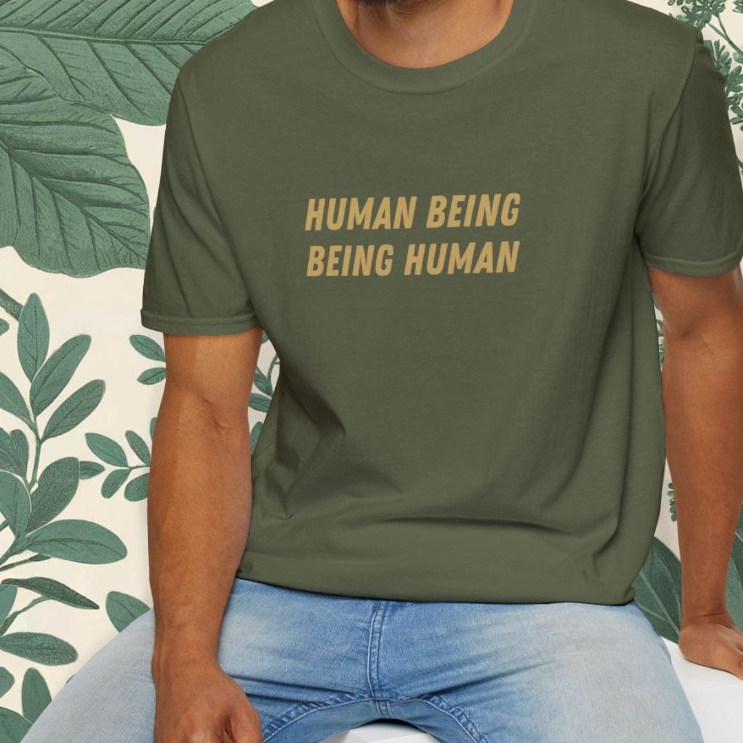Man wearing a green t-shirt with 'HUMAN BEING BEING HUMAN' text, sitting on a white stool against a white background.