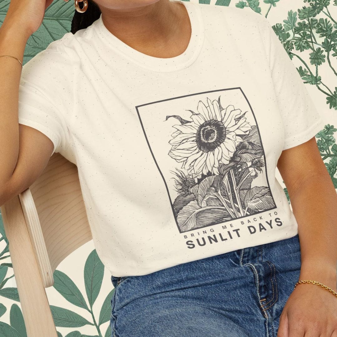 Woman wearing a white t-shirt with a sunflower graphic and 'Sunlit Days' text, sitting on a chair.