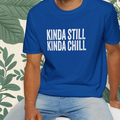 Man wearing a blue t-shirt with 'KINDA STILL KINDA CHILL' text, sitting on a white stool with a green leafy background.