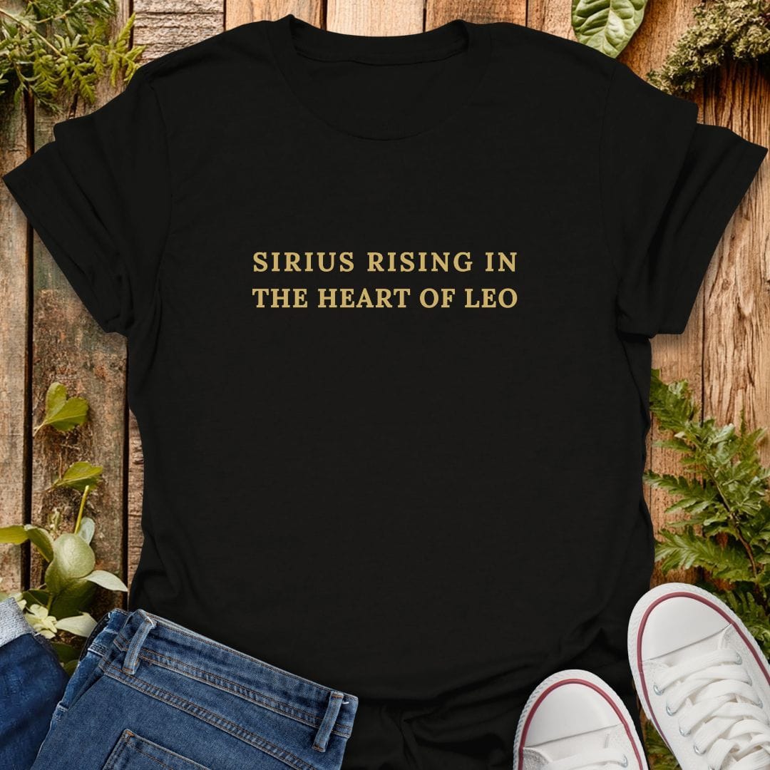 Black short-sleeve T-shirt laid flat on a rustic wooden surface with green plants and white sneakers as styling props. The shirt has gold text reading 'SIRIUS RISING IN THE HEART OF LEO'.
