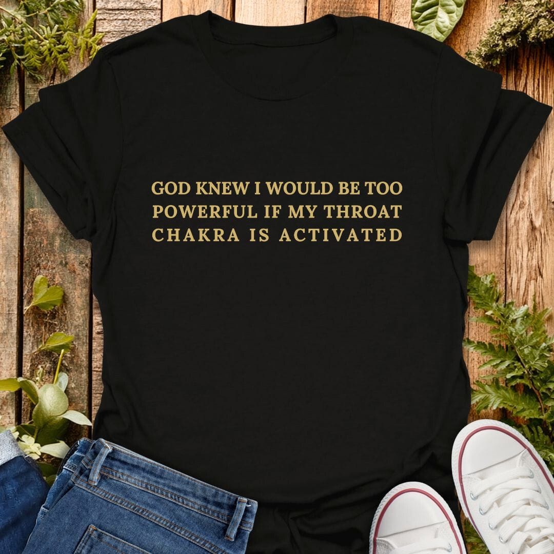 Black t-shirt with "God knew I would be too powerful if my throat chakra is activated" text on a wooden background with plants, jeans, and sneakers.
