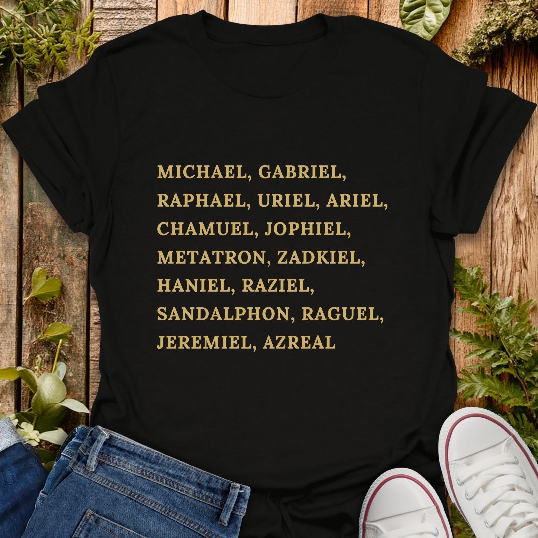 A black short-sleeve T-shirt laid flat on a rustic wooden surface with greenery and jeans, with a pair of white sneakers partly visible at the bottom right. The shirt has a list of names of archangels printed in gold text of on the front.