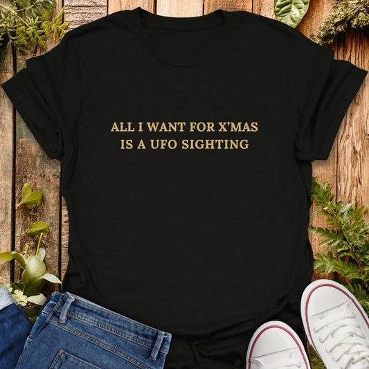 Black t-shirt with 'All I want for x'mas is a UFO sighting' text on a wooden surface with plants, jeans and sneakers.