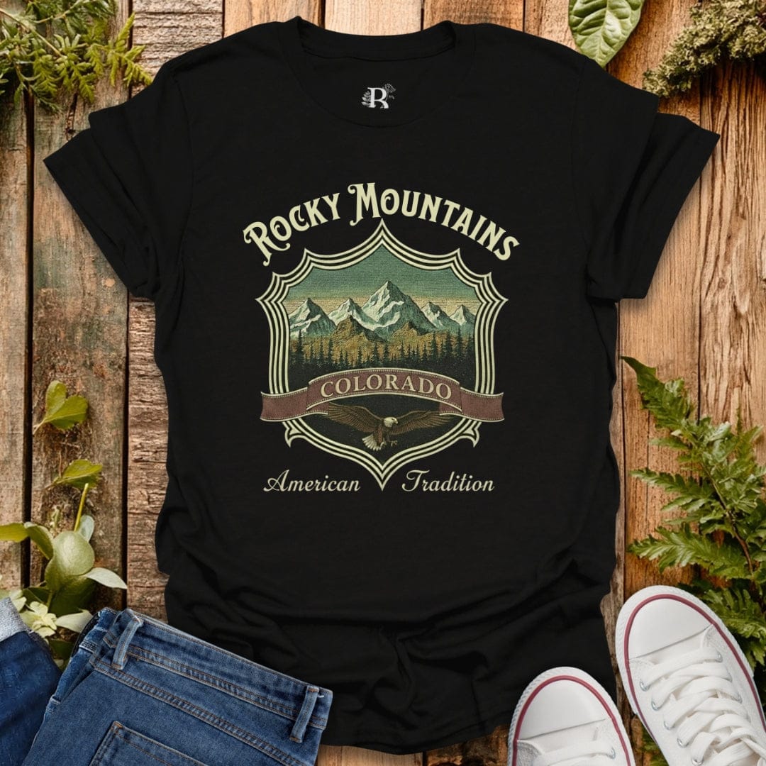Black t-shirt with 'Rocky Mountains Colorado American Tradition' design on a wooden surface with jeans and sneakers.