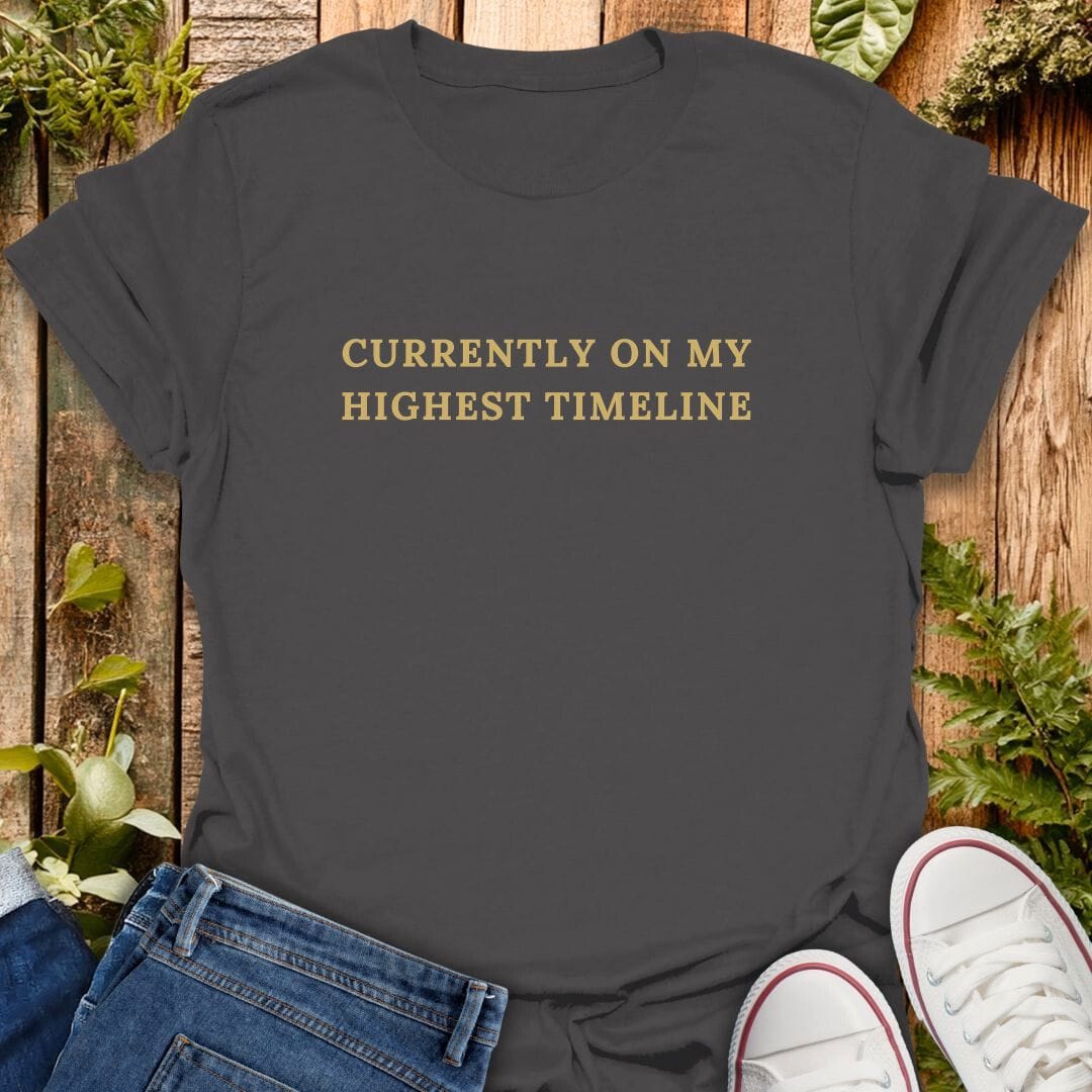 Grey t-shirt with 'Currently on my highest timeline' text on a wooden surface with plants, jeans and sneakers.