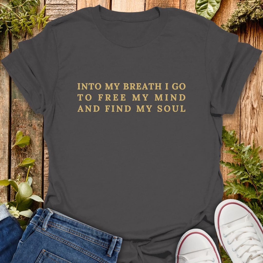 Grey t-shirt with gold text 'Into My Breath I Go To Free My Mind And Find My Soul' on a wooden surface with plants, sneakers and jeans.