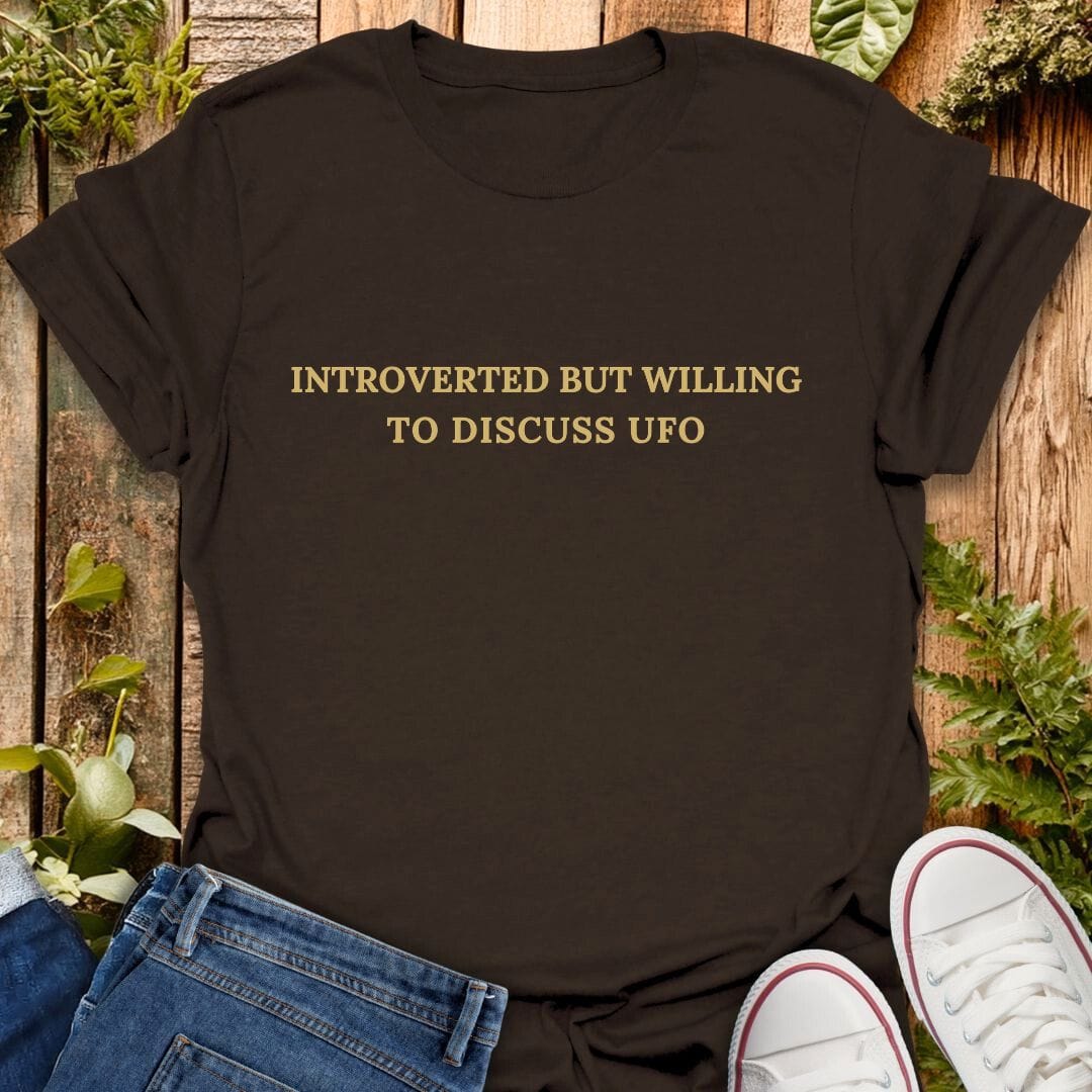 Brown t-shirt with 'Introverted but willing to discuss UFO' text on a wooden background with plants, jeans and sneakers.