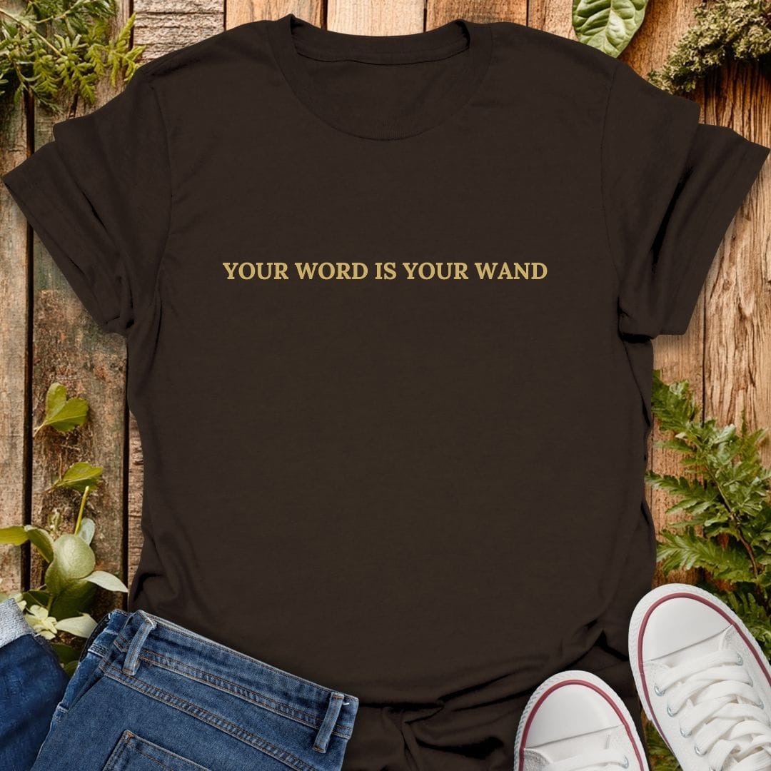 A dark brown short-sleeve t-shirt with the phrase 'YOUR WORD IS YOUR WAND' printed in gold lettering across the chest. The shirt is styled in a flat-lay arrangement with blue jeans, white sneakers, and green foliage on a rustic wooden background.