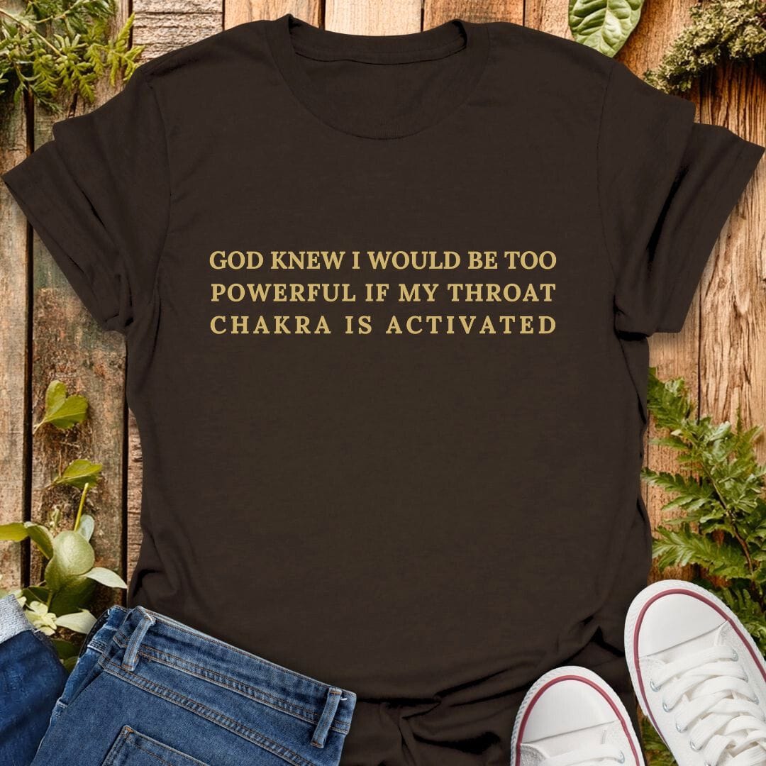 Brown t-shirt with "God knew I would be too powerful if my throat chakra is activated" text on a wooden background with plants, jeans, and sneakers.