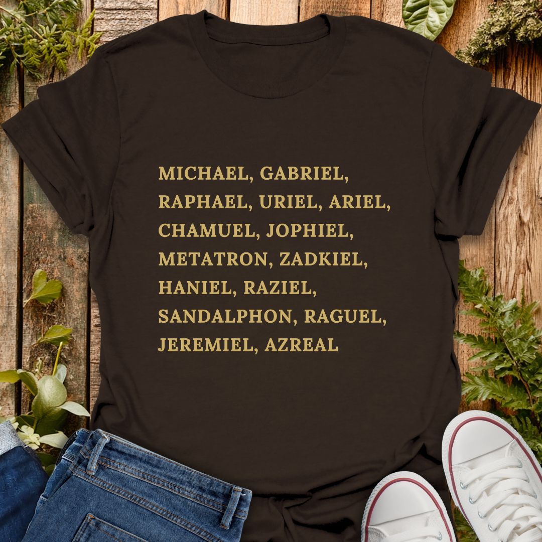 Brown short-sleeve T-shirt laid flat on a rustic wooden surface with greenery and jeans, with a pair of white sneakers partly visible at the bottom right. The shirt has a list of names of archangels printed in gold text of on the front.