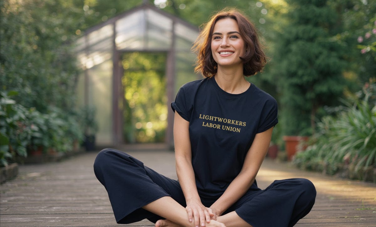 Woman sitting outdoors wearing a black t-shirt with text "lightworkers labor union" surrounded by greenery.