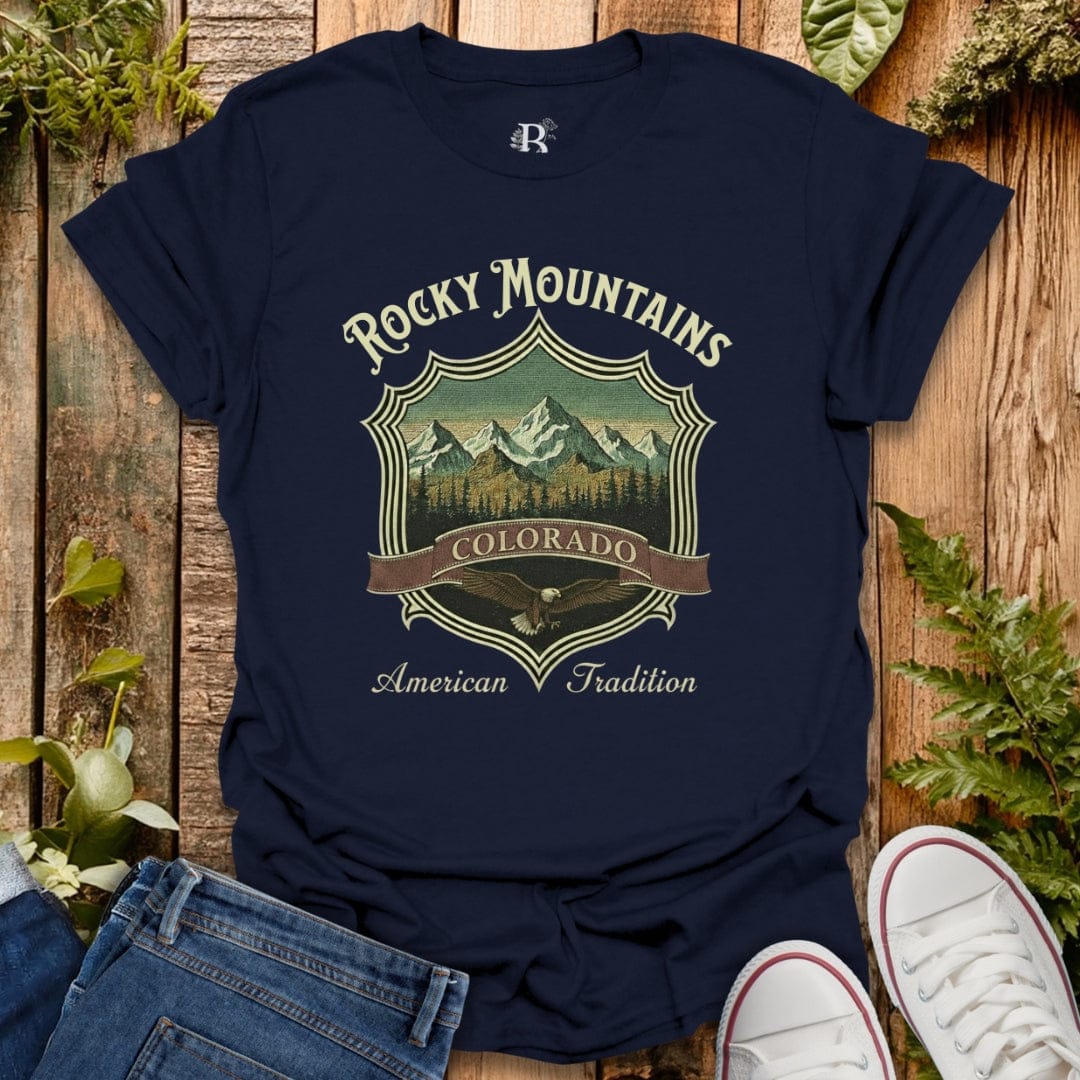 Navy blue t-shirt with 'Rocky Mountains Colorado American Tradition' design on a wooden surface with jeans and white sneakers.