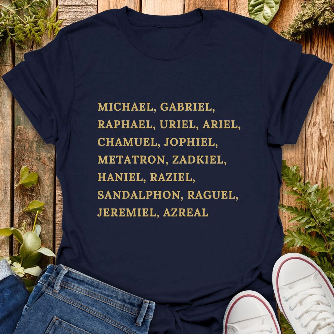A navy blue short-sleeve T-shirt laid flat on a rustic wooden surface with greenery and jeans, with a pair of white sneakers partly visible at the bottom right. The shirt has a list of names of archangels printed in gold text of on the front.