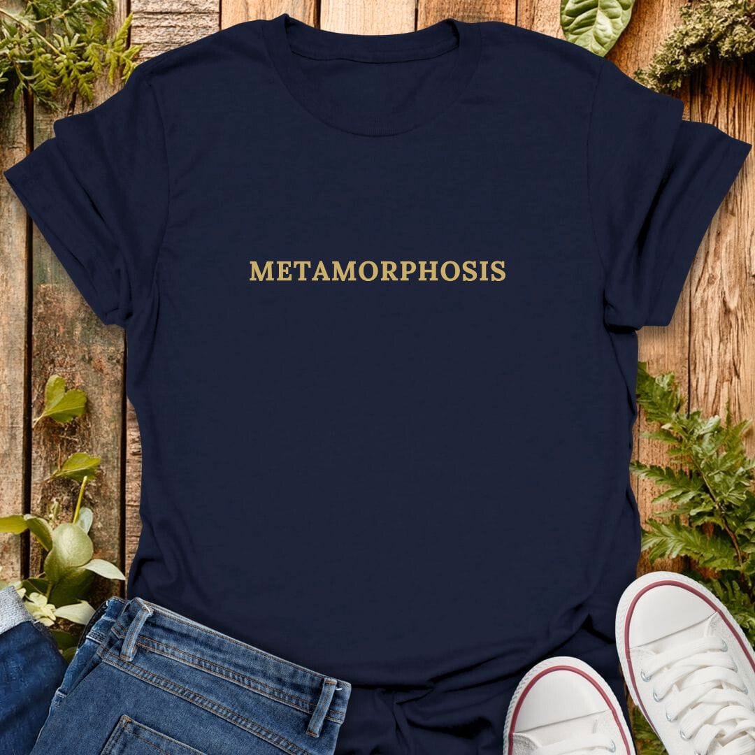 Navy blue t-shirt with 'Metamorphosis' text on a wooden surface with plants, jeans and sneakers.