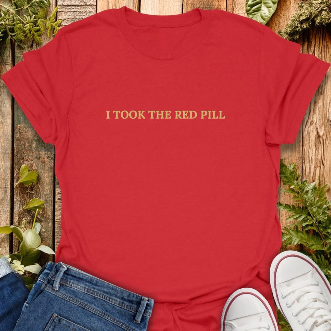 Red t-shirt with 'I took the red pill' text on a wooden surface with plants, jeans and sneakers.